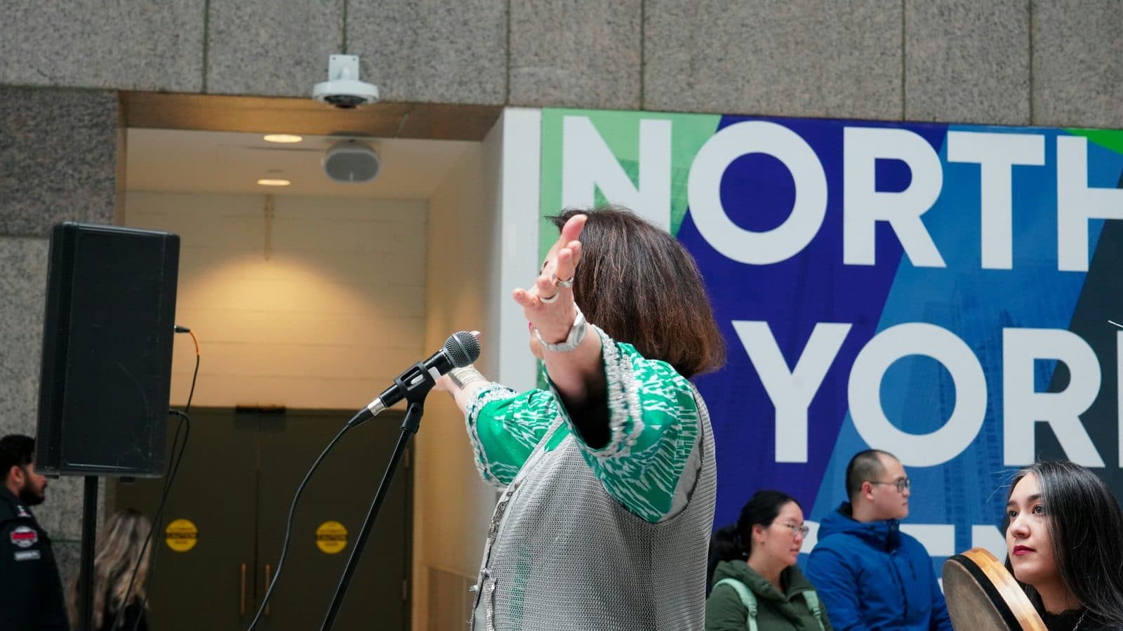 A host speaking into a microphone with an audience behind her, like a quiz master reading out the next question